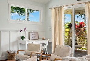 A room with a desk, two chairs, a window with a view of palm trees, and a door leading to an outdoor area with plants.
