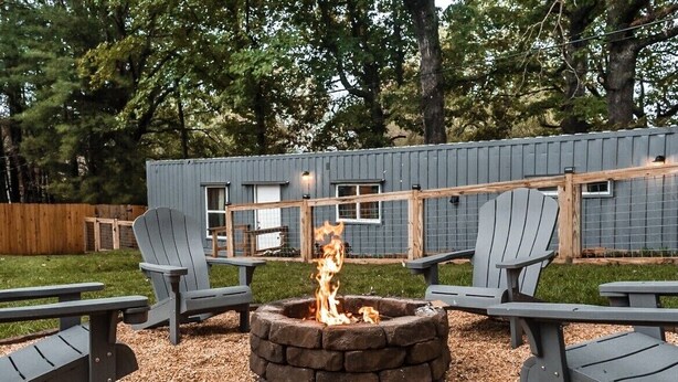 A fire pit surrounded by Adirondack chairs in a backyard with a wooden fence and a building in the background. - Container Home w/ a Fenced Yard (Hendersonville)