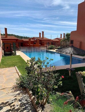 A swimming pool surrounded by buildings with red-tiled roofs, a clear sky, and some greenery.