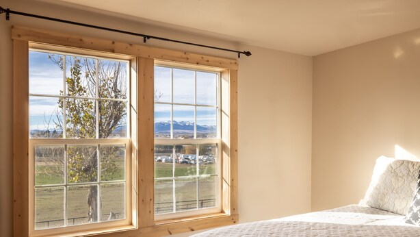 Ein Schlafzimmer mit großem Fenster, einem Bett mit gemustertem Überwurf und Blick auf eine Landschaft mit Bergen und einem Feld.