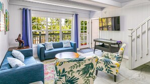 Una sala de estar con un sofá azul, un sillón floral y una mesa de centro. Hay un televisor montado en la pared y una escalera que sube.