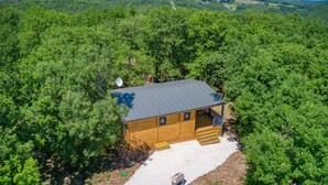 Une cabane en bois, surmontée d’un toit en métal, entourée d’une forêt dense, avec une allée gravillonnée et un petit abri de jardin.