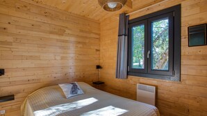 Une chambre dans une cabane en bois, avec un lit, une fenêtre avec des rideaux, une table de chevet et un luminaire fixé au mur.
