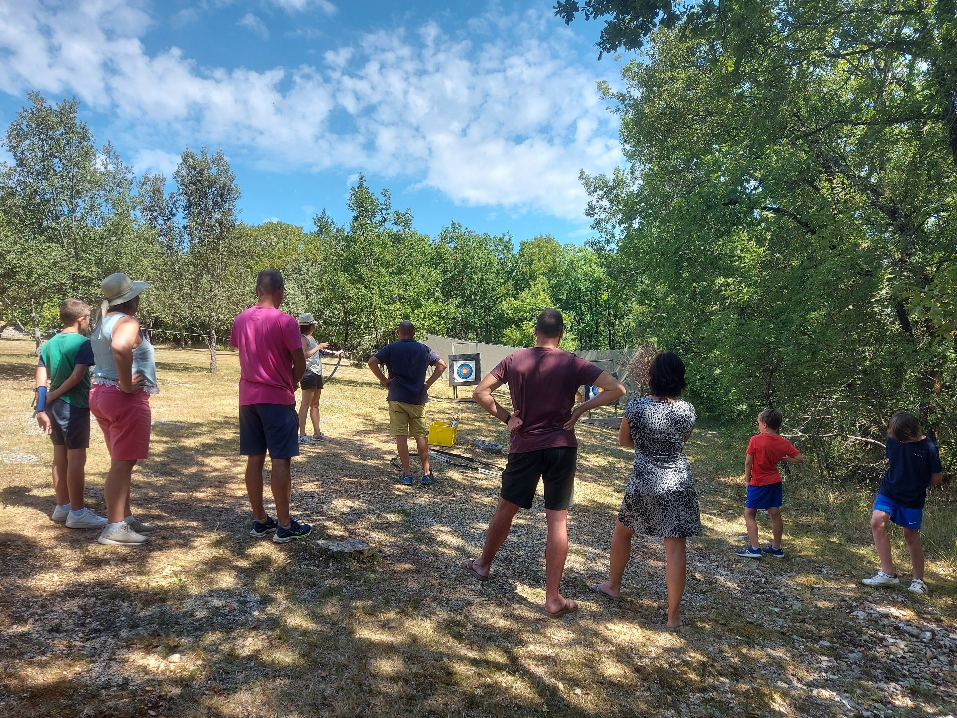 A group of people standing outdoors, possibly at an archery range, with trees and a clear sky in the background.