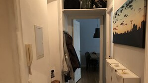 A narrow hallway with wooden flooring, a coat rack, a shoe rack, and a small table with a chair.