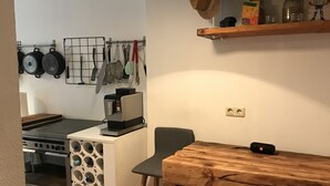 A compact kitchen with a wooden table, a hanging light, and a shelf with bottles.