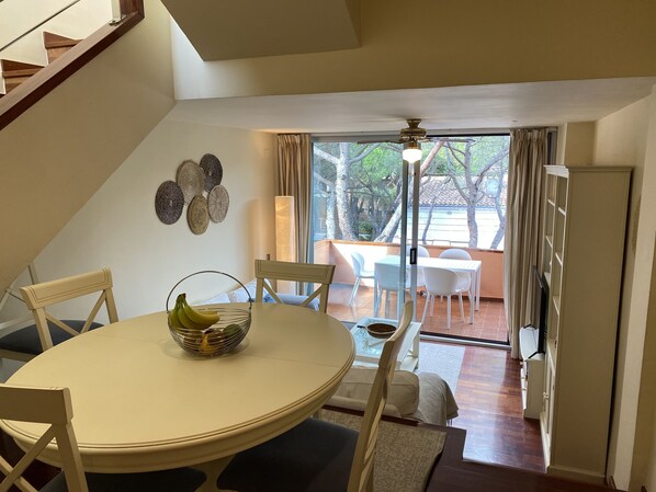 Un comedor con una mesa redonda, sillas y un cesto de plátanos. Se ve un patio exterior con sillas y una mesa.