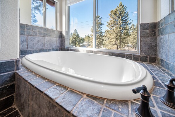 A large, oval-shaped bathtub in a bathroom with stone tile walls and a window providing a view of trees.