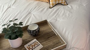 Ein ordentlich bezogenes Bett mit grauem gepolsterten Kopfteil, weißer Bettwäsche und dekorativen Kissen. Auf dem Bett steht ein Tablett mit einer Topfpflanze, einer Kerze und einer motivierenden Karte.