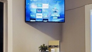 Ein Wohnzimmer mit montiertem Fernseher, der Streaming-Dienste anzeigt, einem kleinen Tisch mit einem 'WELCOME!'-Schild und einem runden Couchtisch mit Dekoration.