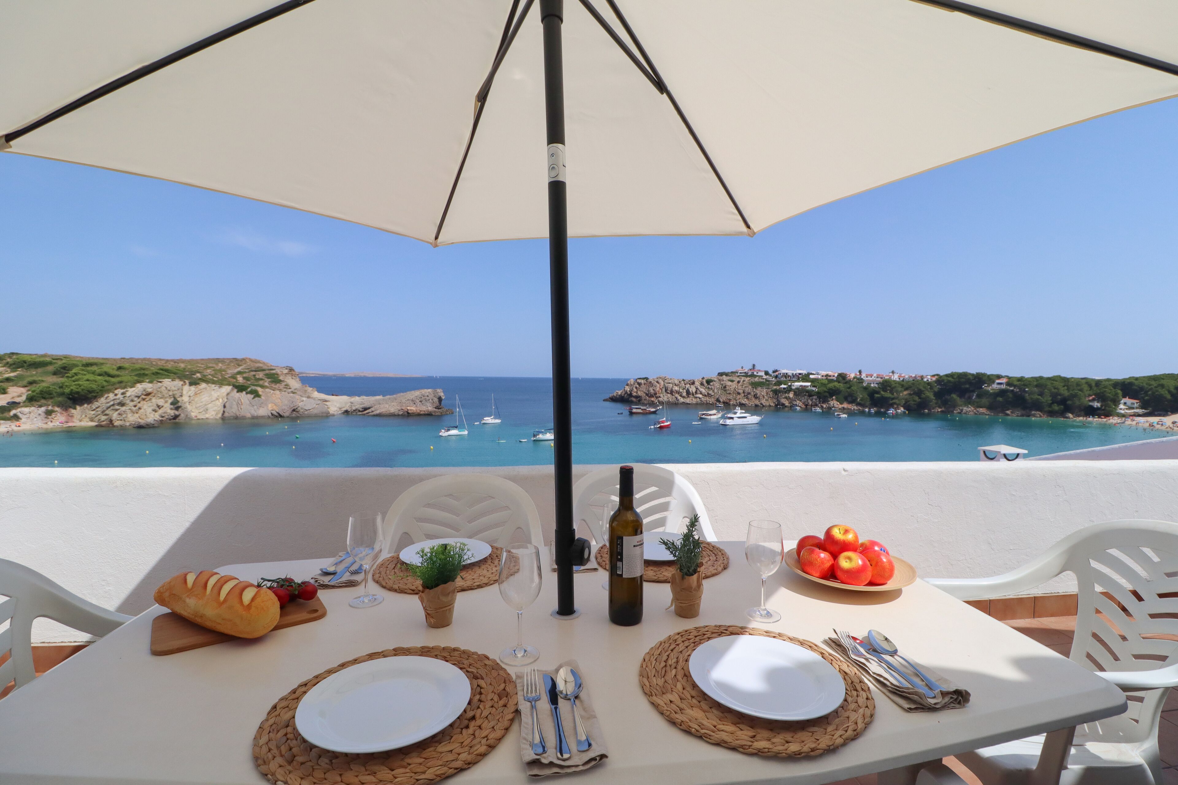 Une table dressée pour deux, avec vue sur la mer et les bateaux, une miche de pain, une bouteille et une assiette de fruits.