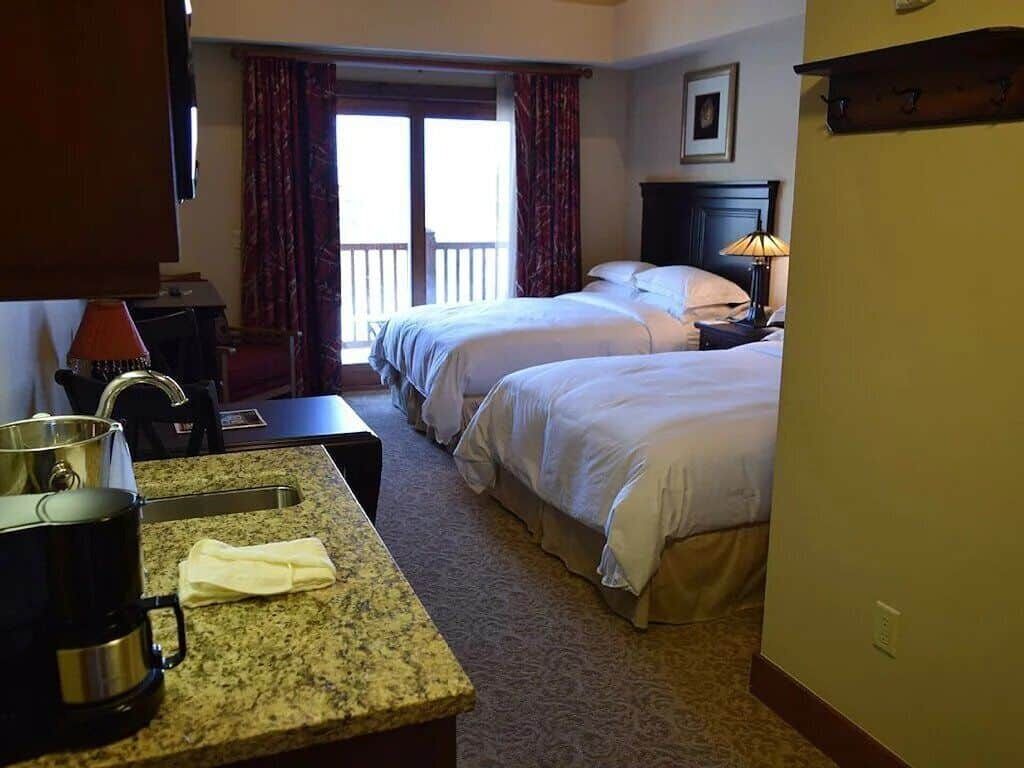 Ein Hotelzimmer mit zwei Betten, einer Küchenzeile mit Kaffeemaschine und einem Balkon mit Vorhängen.