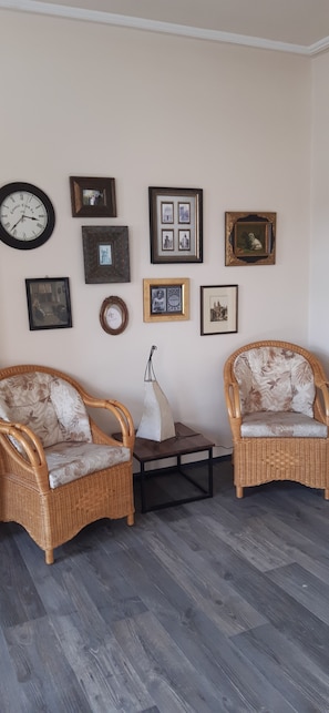 A room with two wicker chairs, a small table, and a wall adorned with various framed pictures.