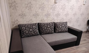 A modern sofa with a grey fabric and patterned cushions, positioned against a patterned wallpaper and a wooden floor.