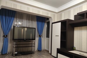 Un salon avec un téléviseur à écran plat posé sur un meuble, des rideaux bleus, un buffet en bois et une porte blanche.