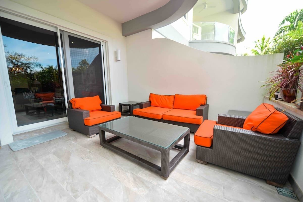 A modern outdoor living area with a glass-top coffee table, orange cushioned sofas, and a dark wicker sofa.