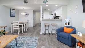 Ein modernes Wohnzimmer mit einem blauen Sofa, eine Essecke mit weißen Stühlen, eine Küche mit weißen Schränken und eine Bar mit Hockern.