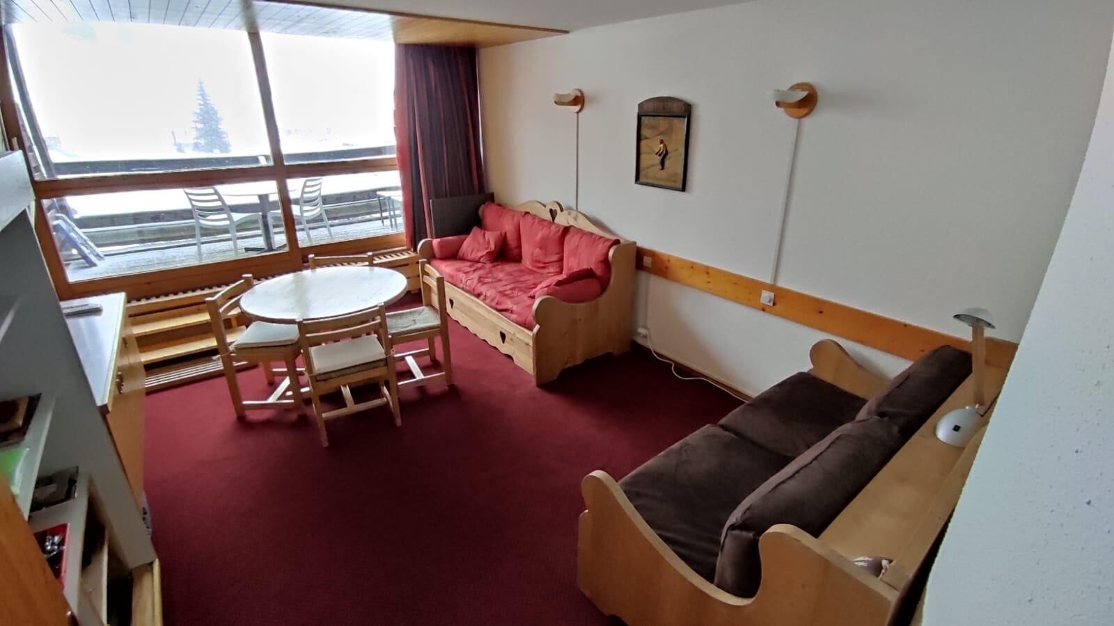 Una habitación pequeña con una mesa y sillas de madera, un sofá rojo y una cama individual.