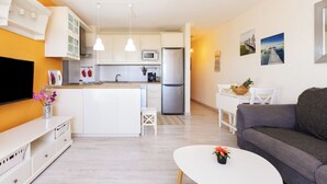 A modern kitchen with white cabinets, a central island, and stainless steel appliances. There is a dining area with a white table and chairs, and a gray sofa in the living room.