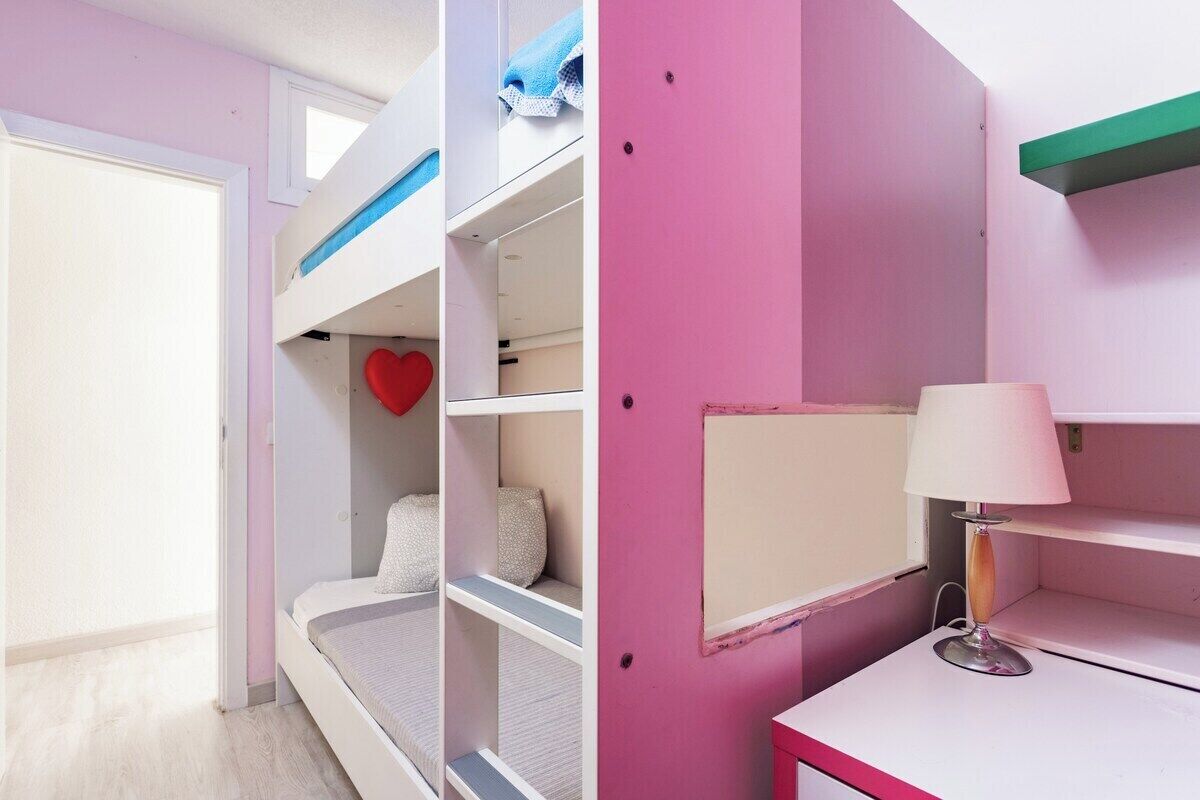 A bunk bed with a red heart decoration, a bedside table with a lamp, and a pink wall with a shelf.