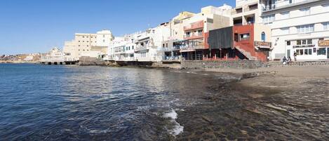 A coastal town with buildings along the waterfront, clear shallow waters, and a rocky beach.