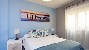 A bedroom with a bed, two bedside tables, a nightstand, a painting of a sunset over a pier, and sheer curtains on a window.