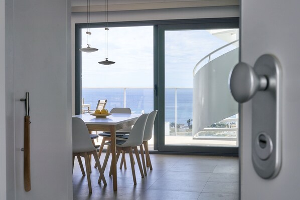A modern dining area with a view of the sea, a wooden dining table, white chairs, and pendant lights.