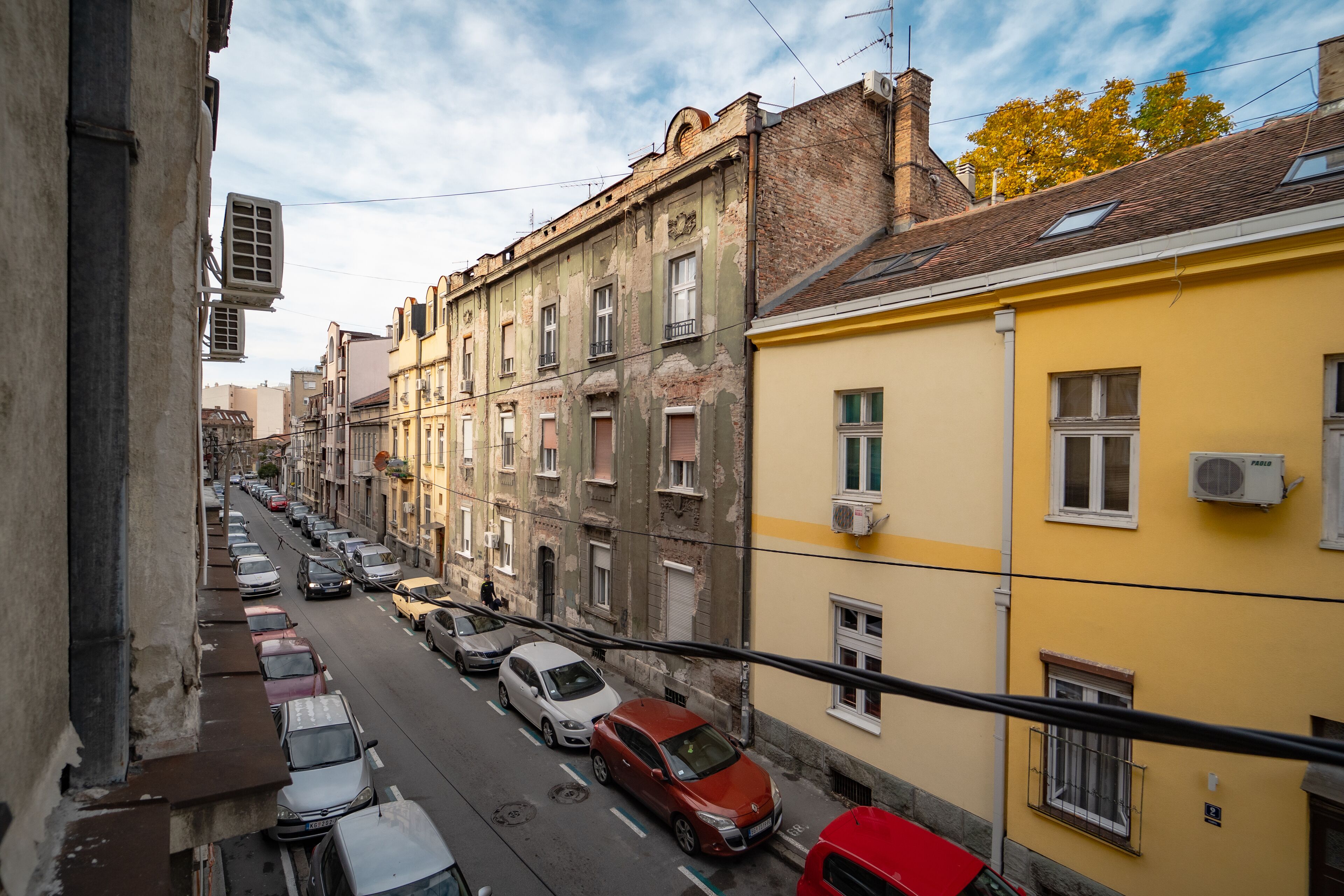 Une rue étroite en milieu urbain, bordée d’immeubles à plusieurs étages, de voitures garées et sous un ciel dégagé.