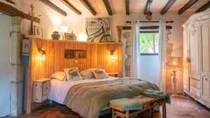 A rustic bedroom with exposed wooden beams, a large bed with a green blanket, a wooden bench, and a stone floor. - COUNTRY HOME (Castelnau-sur-Gupie)