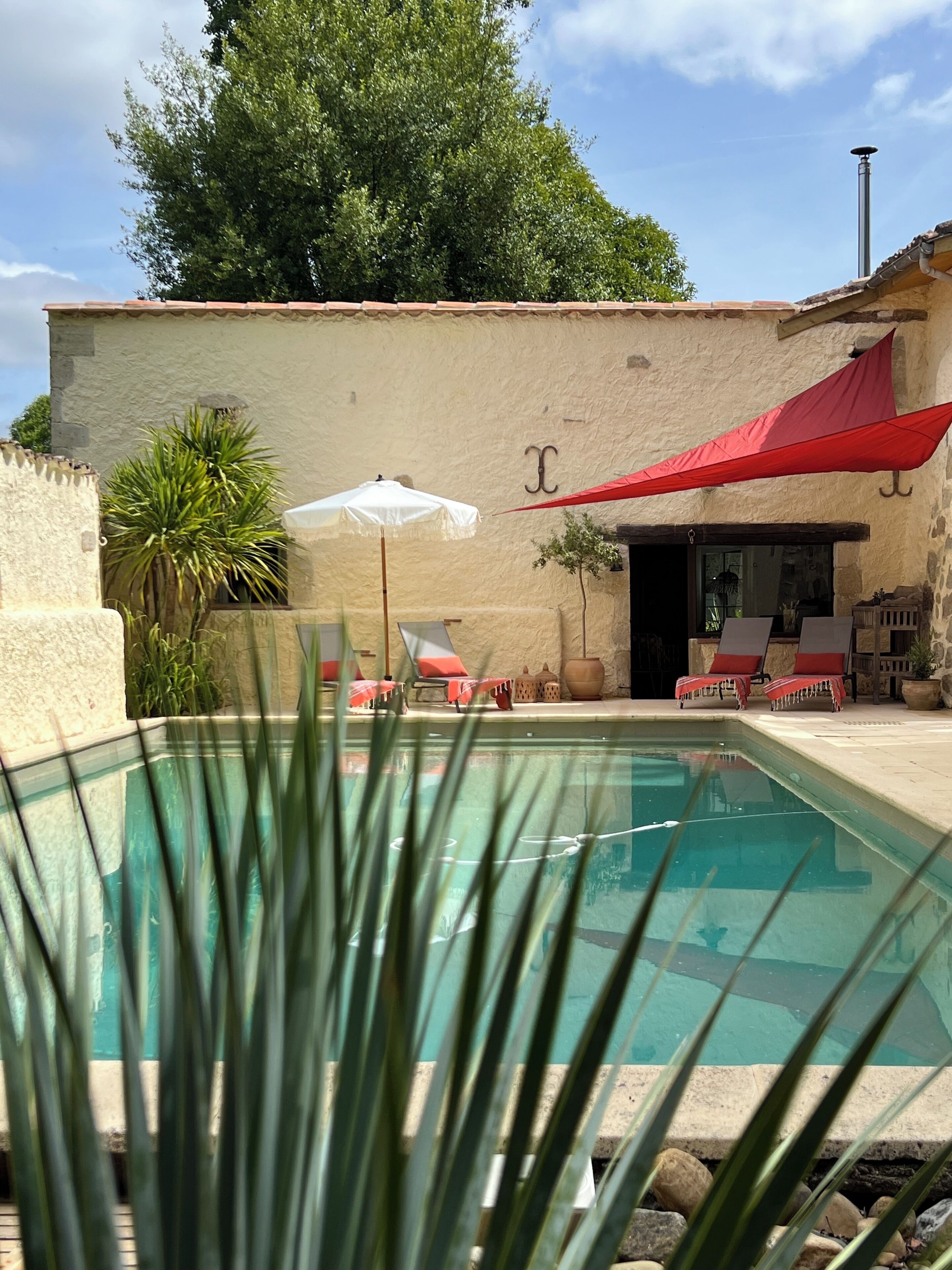 A swimming pool with lounge chairs and a red canopy, surrounded by a stone wall and greenery.