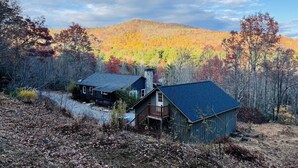 A cabin with a dark roof and a small porch, surrounded by a forest with autumn-colored trees. - The Outpost, on Little Pisgah Mountain - a Secluded Modern Cabin Oasis (Hendersonville)