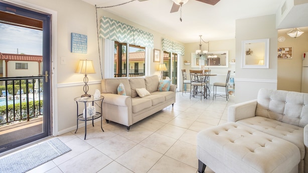 A spacious living room with a beige sofa, a white armchair, and a dining area with a table and chairs. A ceiling fan is visible, and there is a view of a pool through an open door. - Island getaway 3 minutes from sandy beaches. (Treasure Island)