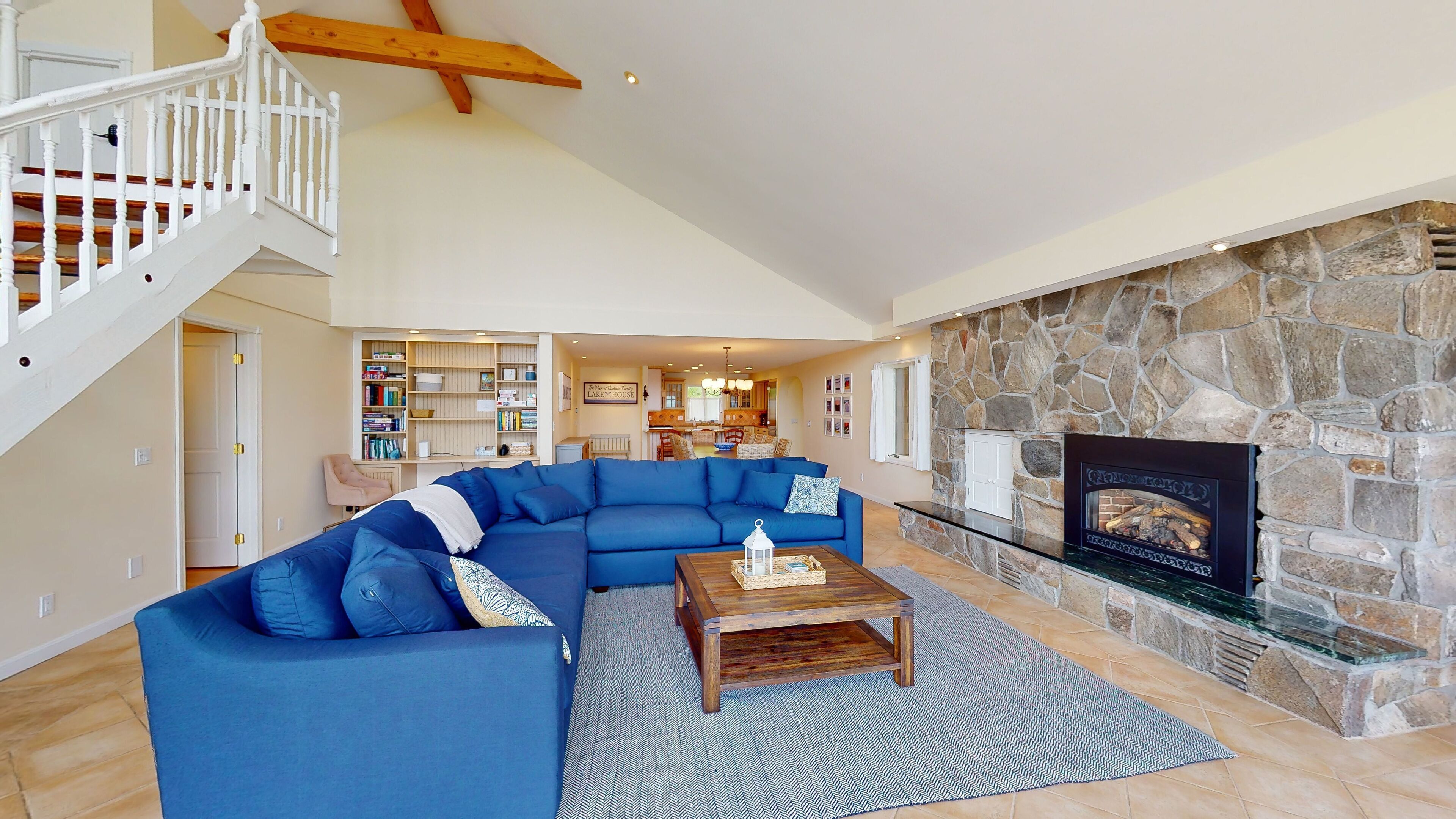 A spacious living room with a blue sectional sofa, a stone fireplace, a wooden coffee table, and a staircase with white railings.