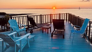 Terraza de madera con sillones de descanso y una mesa, con vista a la puesta de sol sobre el agua.