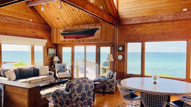 Una cabaña de madera con un ventanal grande que da al mar, amueblada con un sofá, sillas y una mesa de comedor.
