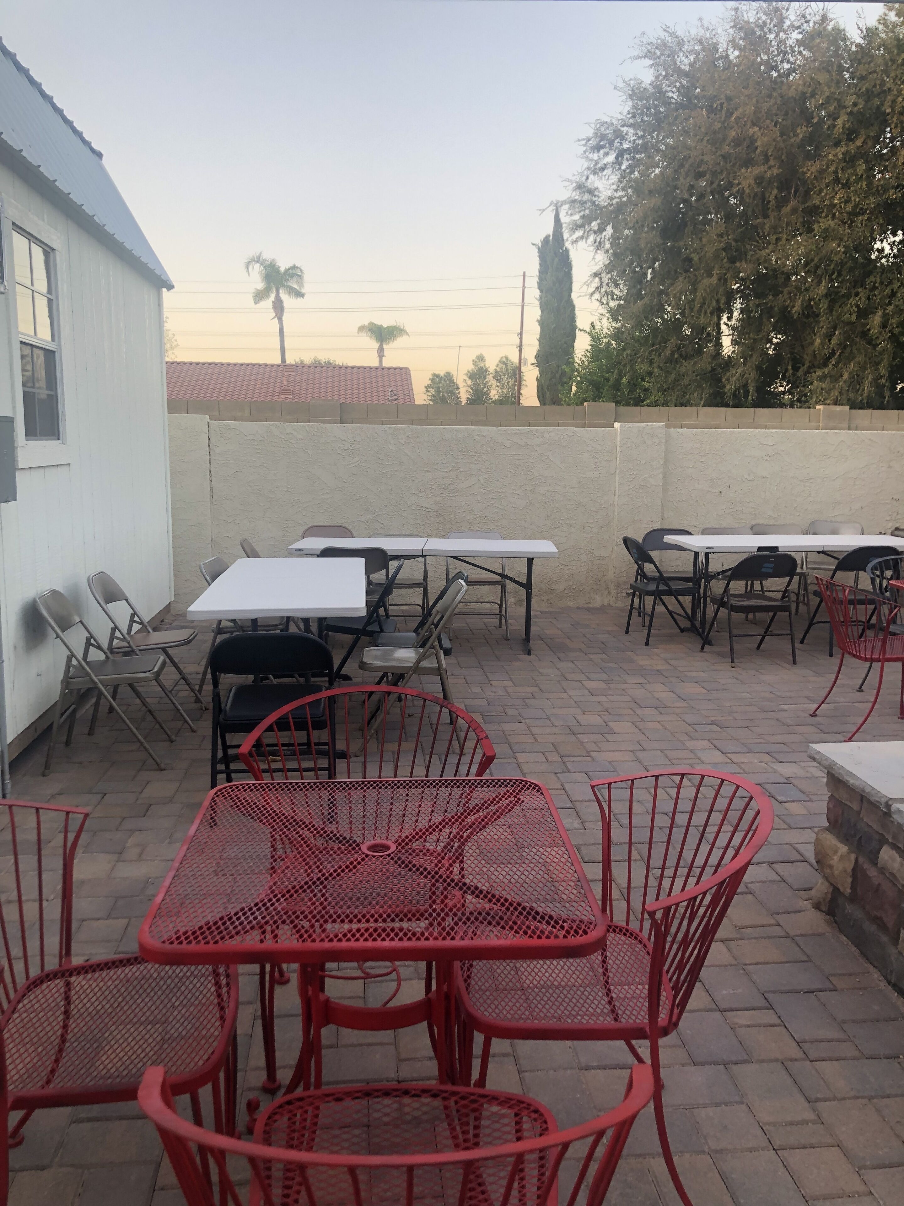 An outdoor patio with tables and chairs, a white building, and trees in the background.
