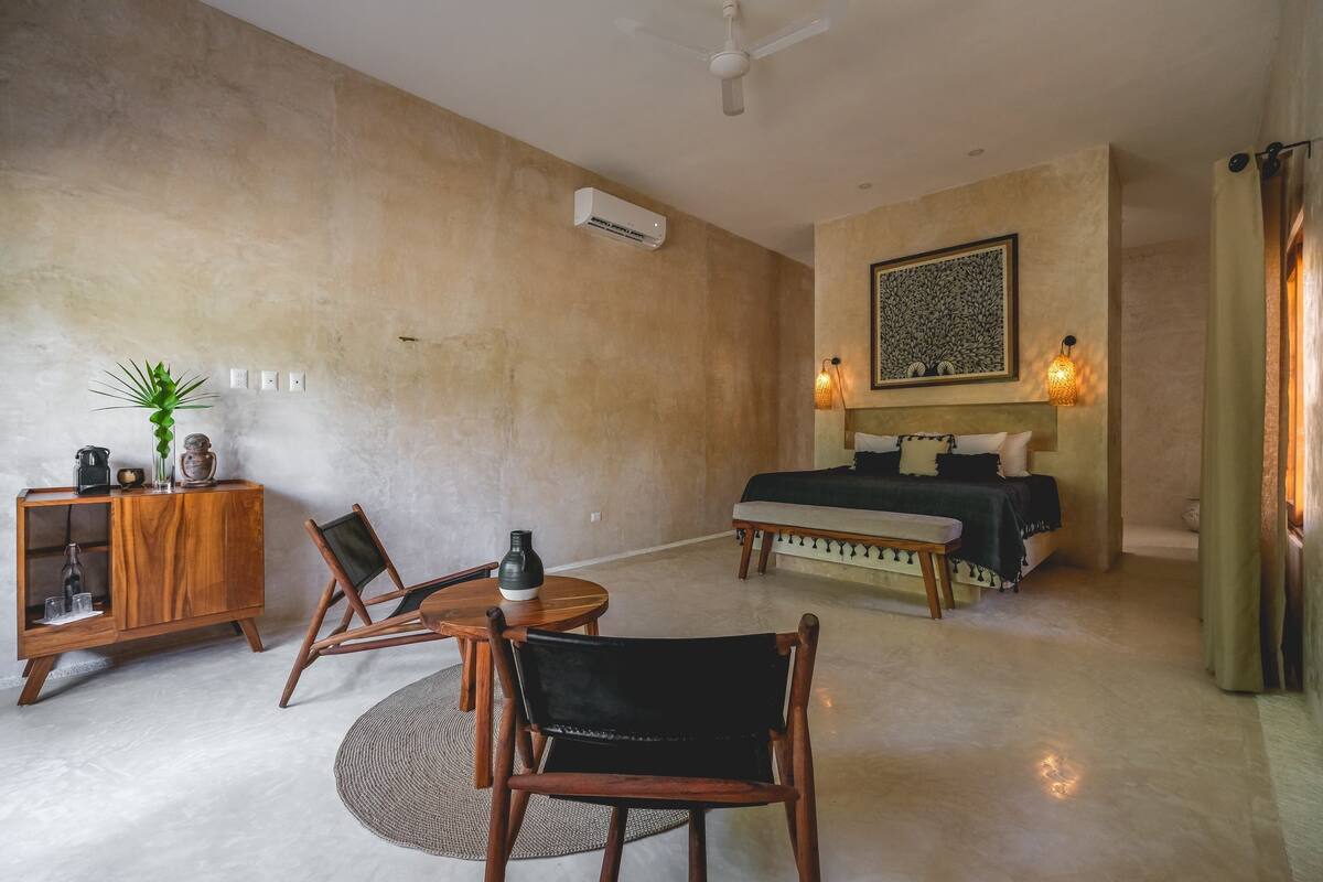 A modern living room with a sofa, a dining table, and a cabinet. There is a bed with a headboard and a wall-mounted air conditioner.