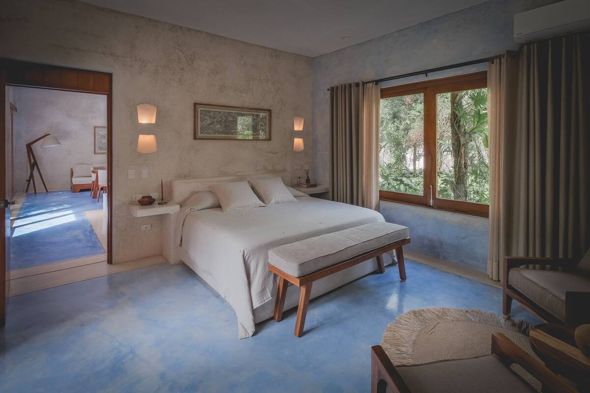 A bedroom with a large bed, bedside lamps, a window with curtains, and a view of greenery outside.