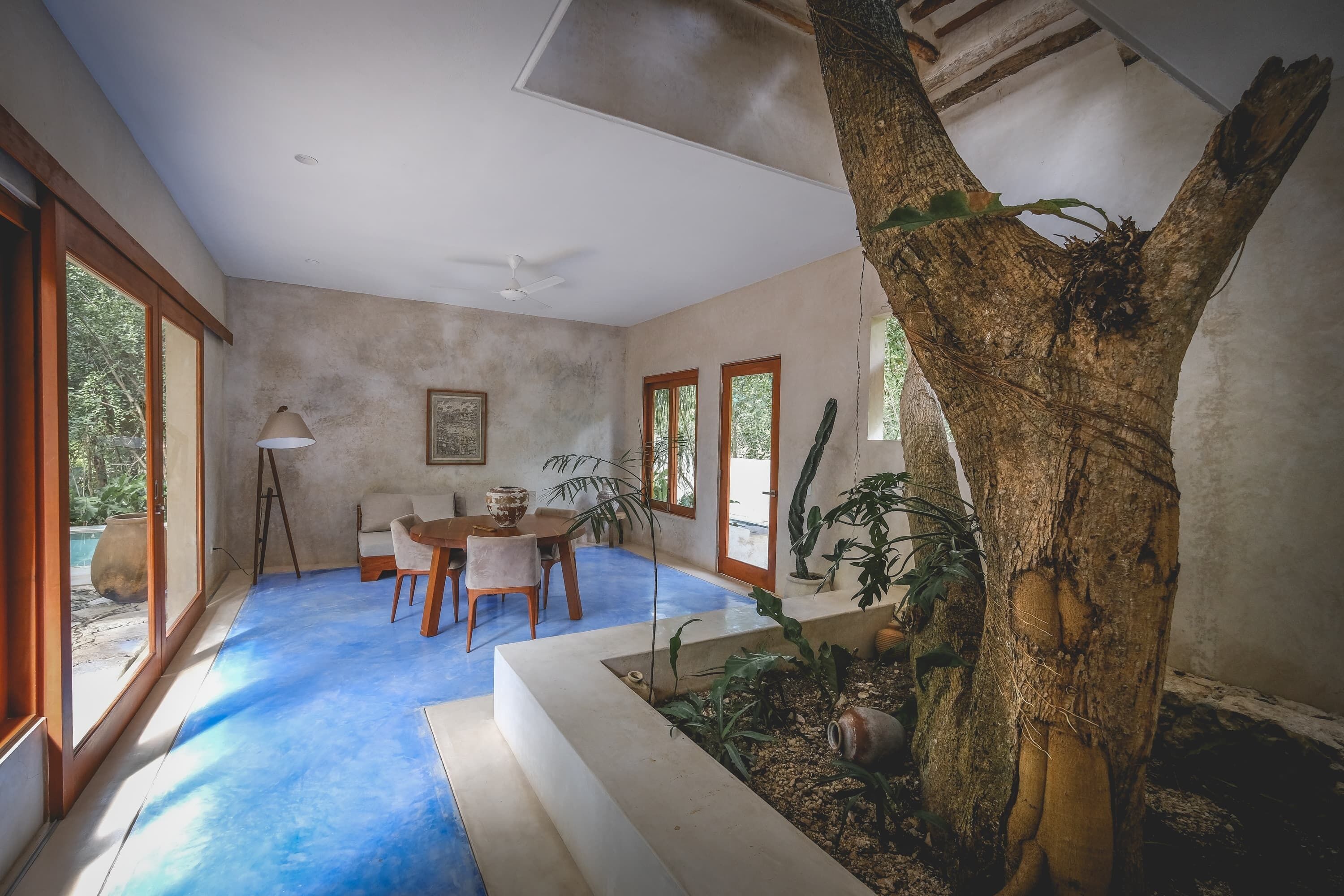 A spacious room with a large window, a ceiling fan, a dining area with a table and chairs, and a tree trunk in the corner.