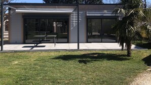 Ein modernes Gebäude mit großen Glasfenstern, einer überdachten Terrasse und einer kleinen Palme im Vordergrund.