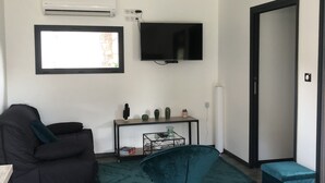 Ein modernes Wohnzimmer mit einer schwarzen Couch, einem petrolfarbenen Sessel, einem Glastisch, einem an der Wand montierten Fernseher und einer Klimaanlage.