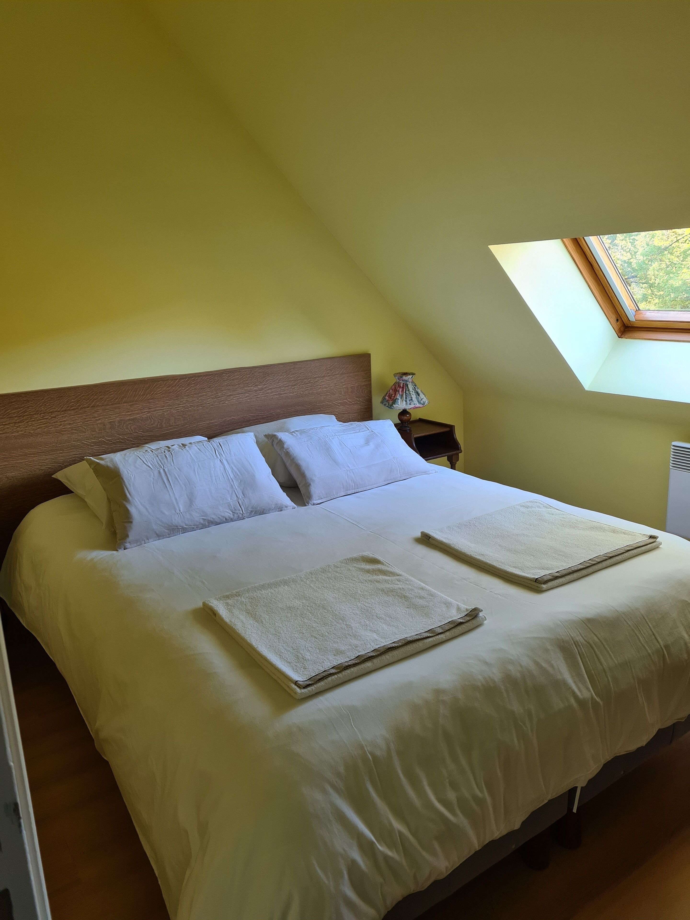 A neatly made bed with white linens and two folded towels, a wooden headboard, a bedside lamp, and a skylight above.
