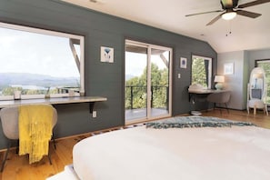 Ein Schlafzimmer mit einem großen Bett, einem Schreibtisch, einem Sessel, einem Balkon mit Blick und einem Deckenventilator.