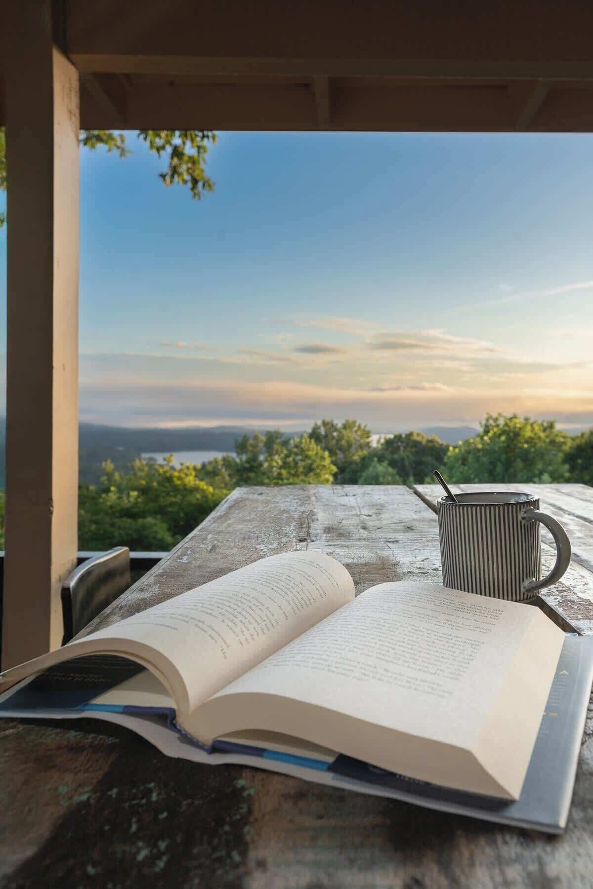 An open book on a wooden surface with a striped mug beside it, under a wooden structure with a view of greenery and a distant landscape.