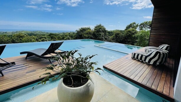 Ein Fernblick-Pool mit Liegestühlen und einer Holzterrasse, umgeben von üppiger Vegetation und blauem Himmel.