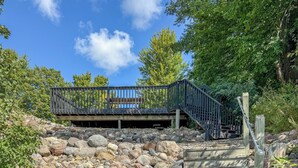 Eine hölzerne Treppe führt zu einem Holzdeck mit Metallgeländer, umgeben von großen Steinen und Grünpflanzen.