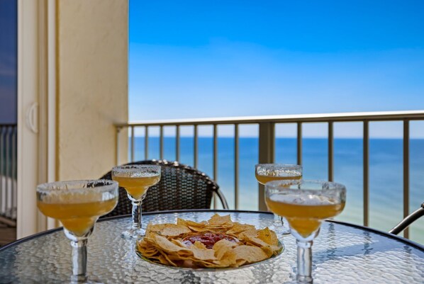 A table with a plate of nachos and three glasses of a yellow beverage, set on a balcony with a view of the ocean.
