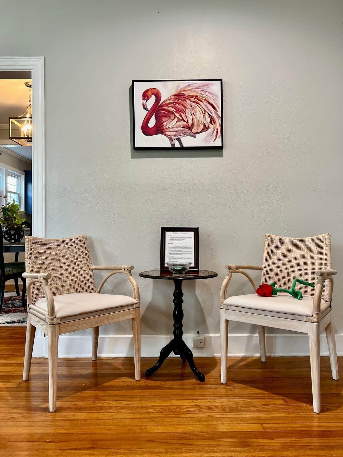 A room with two wicker chairs, a small table with a framed certificate, and a wall-mounted flamingo artwork.
