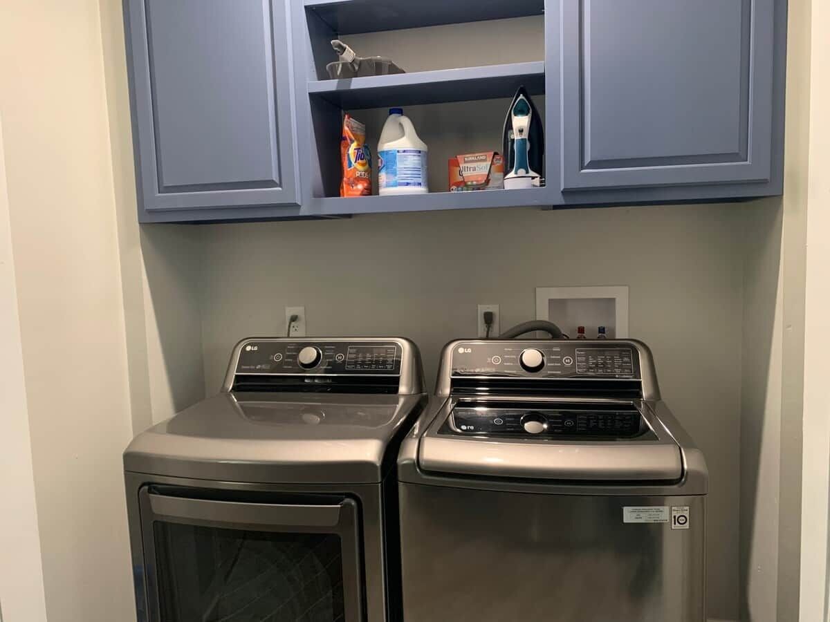 A laundry room with two stacked washers and dryers, a shelf above containing cleaning supplies, and a power outlet on the wall.