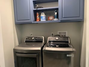 A laundry room with two stacked washers and dryers, a shelf above containing cleaning supplies, and a power outlet on the wall.